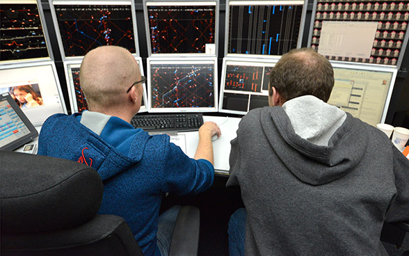 Zwei Männer sitzen vor 10 Monitoren auf denen vor allem rote und blaue Linien zu sehen sind.